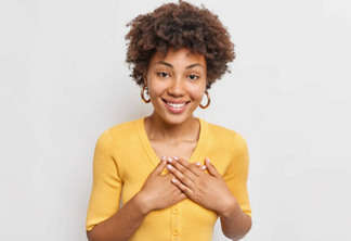 Thank you. Pleased smiling African American woman presses hands to chest expresses gratitude feels touches wears earrings comfortable yellow jumper isolated over white wall. Appreciation concept